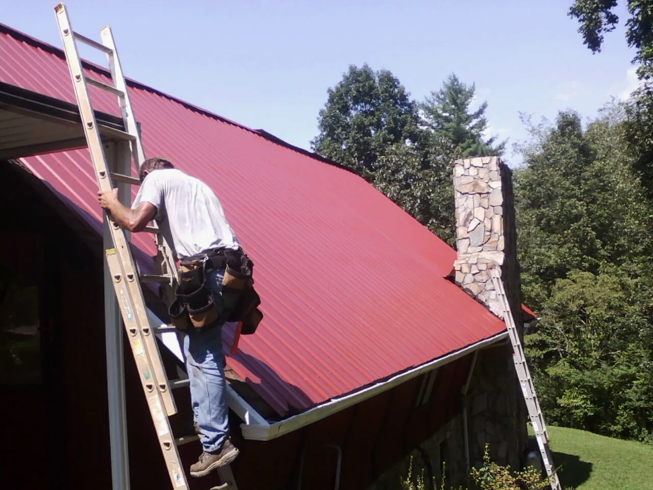 Roofer on a ladder beside a red metal roof and stone chimney — Soffit & Fascia Repair by Roofing Brothers in Roseboro